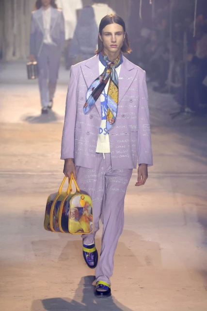 Male fashion model wearing a lavender suit and carrying a duffel bag on a runway during a fashion show.