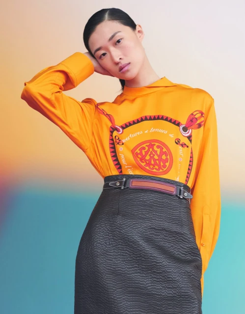 Portrait of an Asian woman wearing a printed orange blouse and a black skirt. She is posing in front of a colorful gradient backdrop.