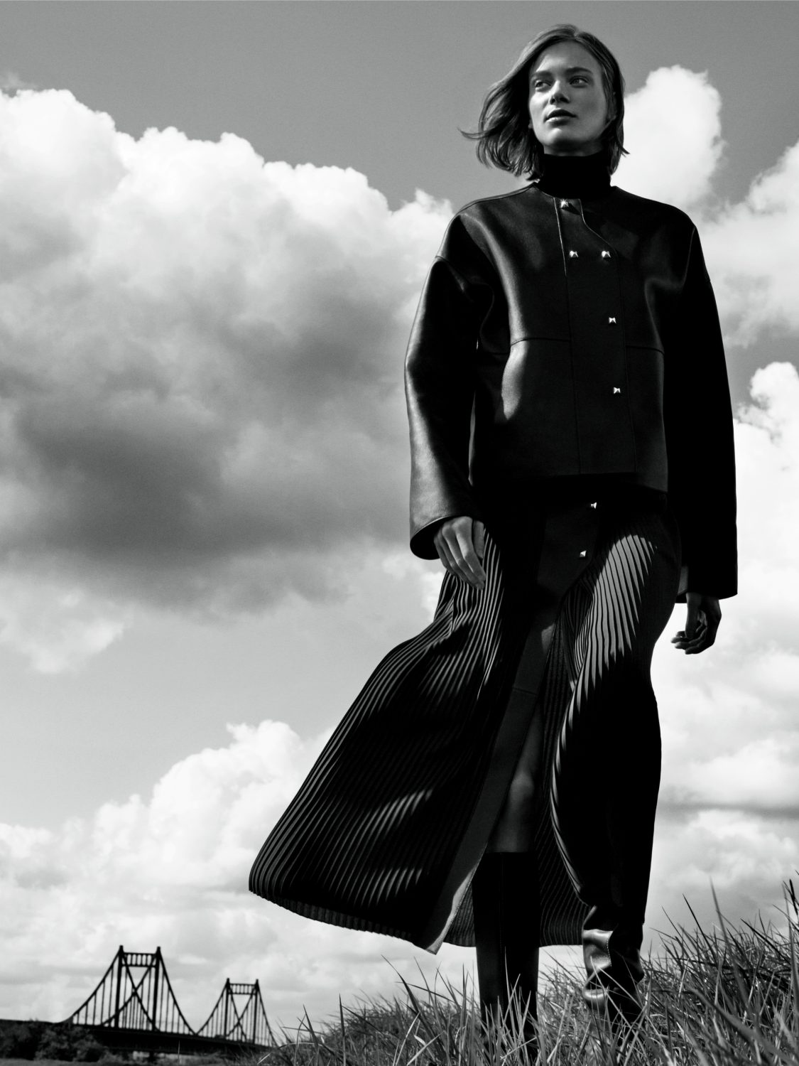 Monochrome fashion portrait of a woman wearing a dark leather jacket and pleated skirt outdoors under a cloudy sky.