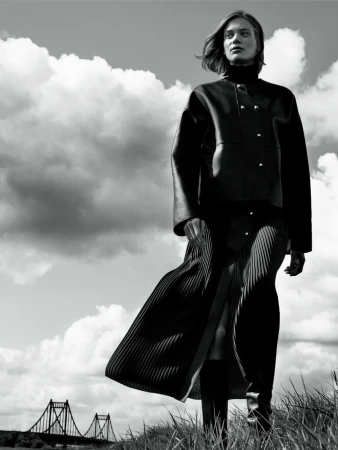 Monochrome fashion portrait of a woman wearing a dark leather jacket and pleated skirt outdoors under a cloudy sky.