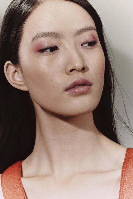 Close-up portrait of a young Asian woman with pink eye shadow and glossy lips, shot with shallow depth of field in a photo studio.
