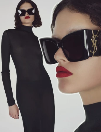 Close-up portrait of a woman wearing oversized black sunglasses with gold YSL logo and bright red lipstick against a white background.