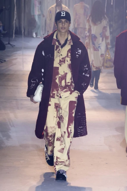 Male model walks the runway wearing a beige patterned shirt and pants with a long maroon coat, accessorized with a baseball cap and white purse.