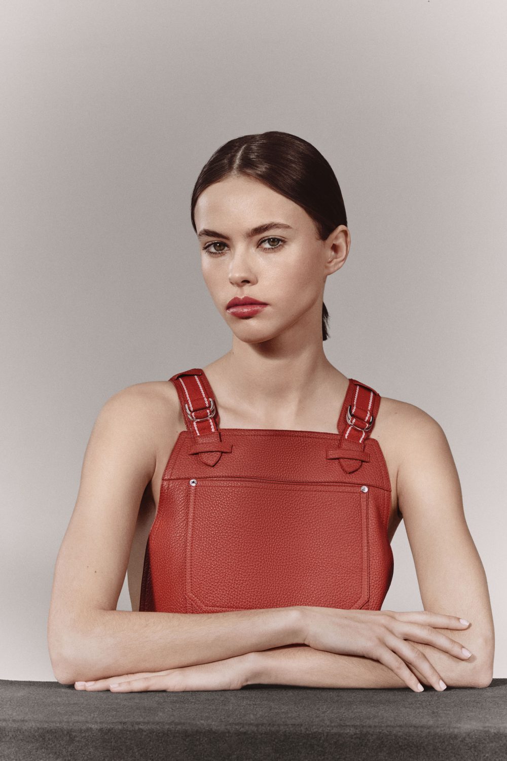 Fashion portrait of a woman in a red overall dress with her arms crossed, set against a neutral backdrop in a studio setting.