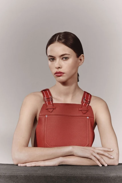 Fashion portrait of a woman in a red overall dress with her arms crossed, set against a neutral backdrop in a studio setting.