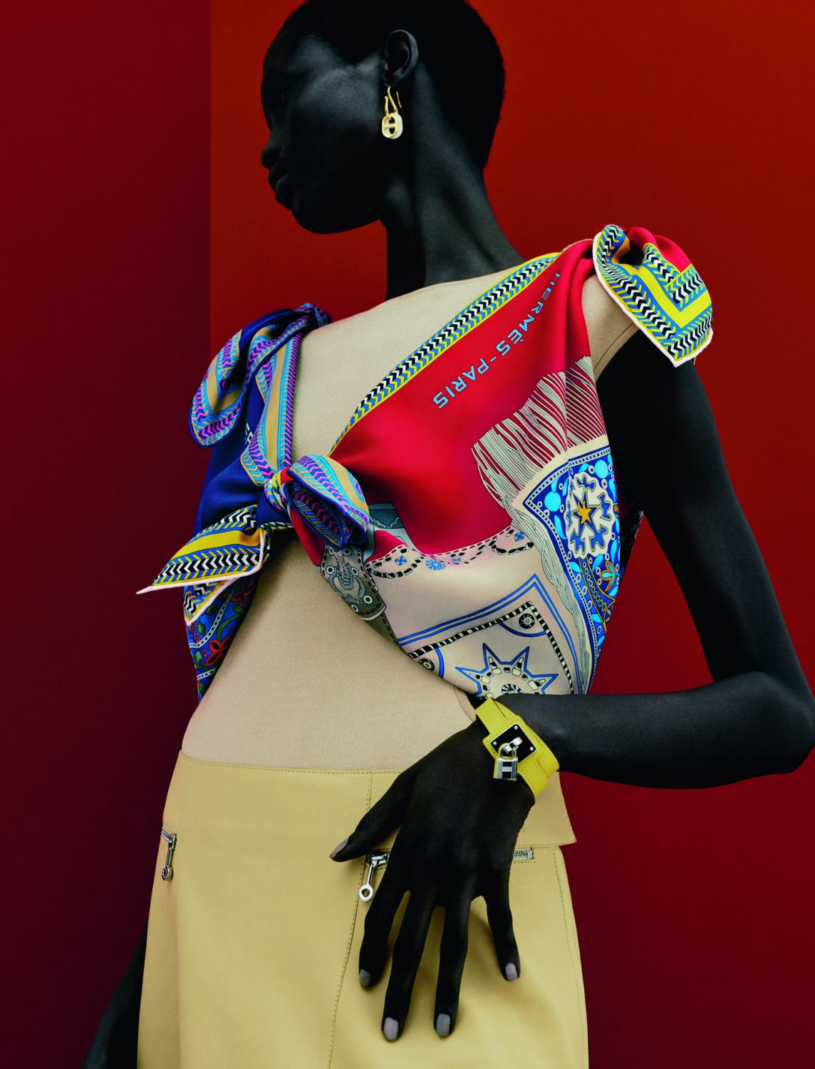 Fashion editorial featuring a Black woman wearing a tan skirt, colorful scarf top, and gold earrings against a gradient red backdrop.