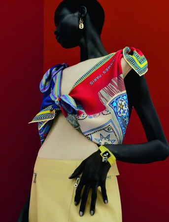 Fashion editorial featuring a Black woman wearing a tan skirt, colorful scarf top, and gold earrings against a gradient red backdrop.
