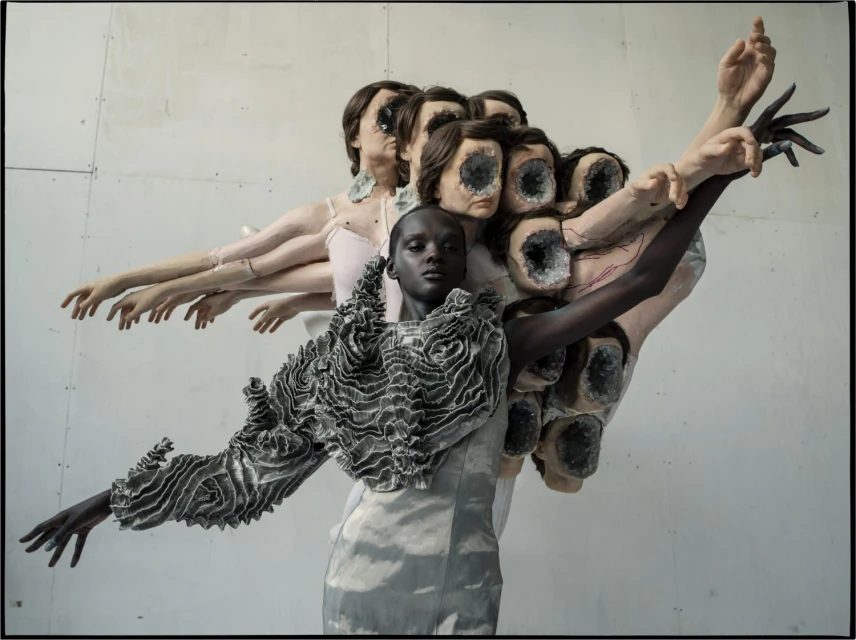 Artistic fashion shot featuring a dark-skinned model in a ruffled top, surrounded by mannequin parts and set against a plain studio backdrop.