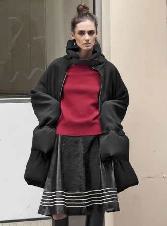 Model with hair in top knot wearing oversized black coat, red sweater, and dark flared skirt standing outdoors