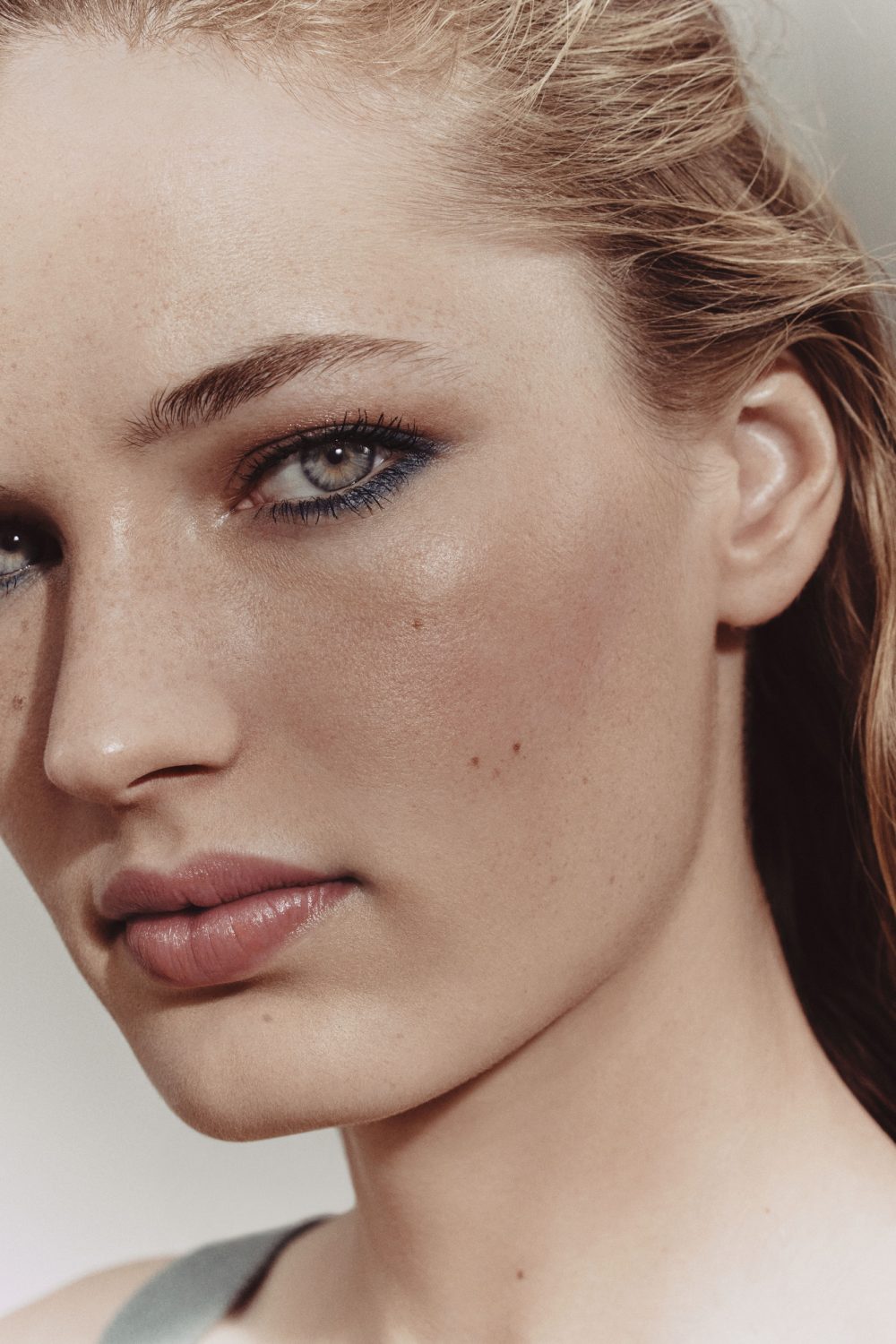 Close-up studio portrait of a young adult woman with fair skin, blue eyes, light brown hair, and natural makeup. Beauty shot, clean aesthetic.