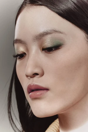 Close-up portrait of a woman wearing shimmering green eyeshadow and glossy lips with soft lighting against a neutral background
