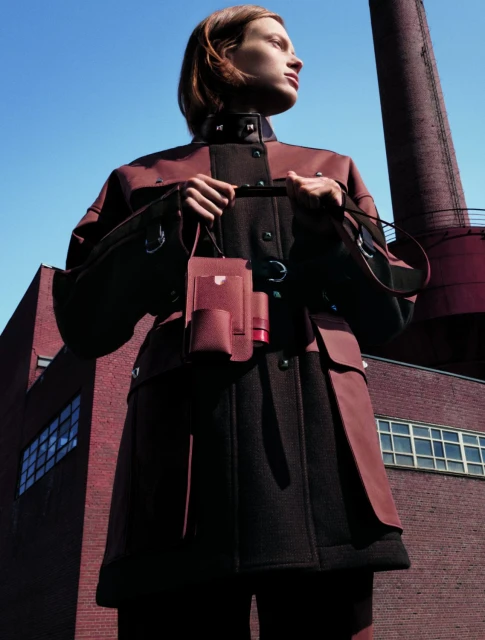 Woman in a burgundy coat stands outdoors in front of a brick building and industrial chimney, holding a burgundy bag.