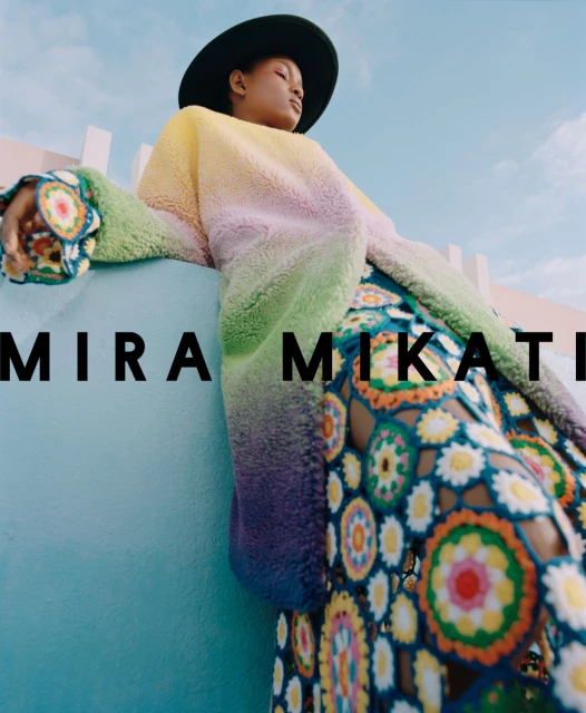 A person in a colorful outfit with a black hat poses against a light blue and white background under a bright sky in a fashion shot.