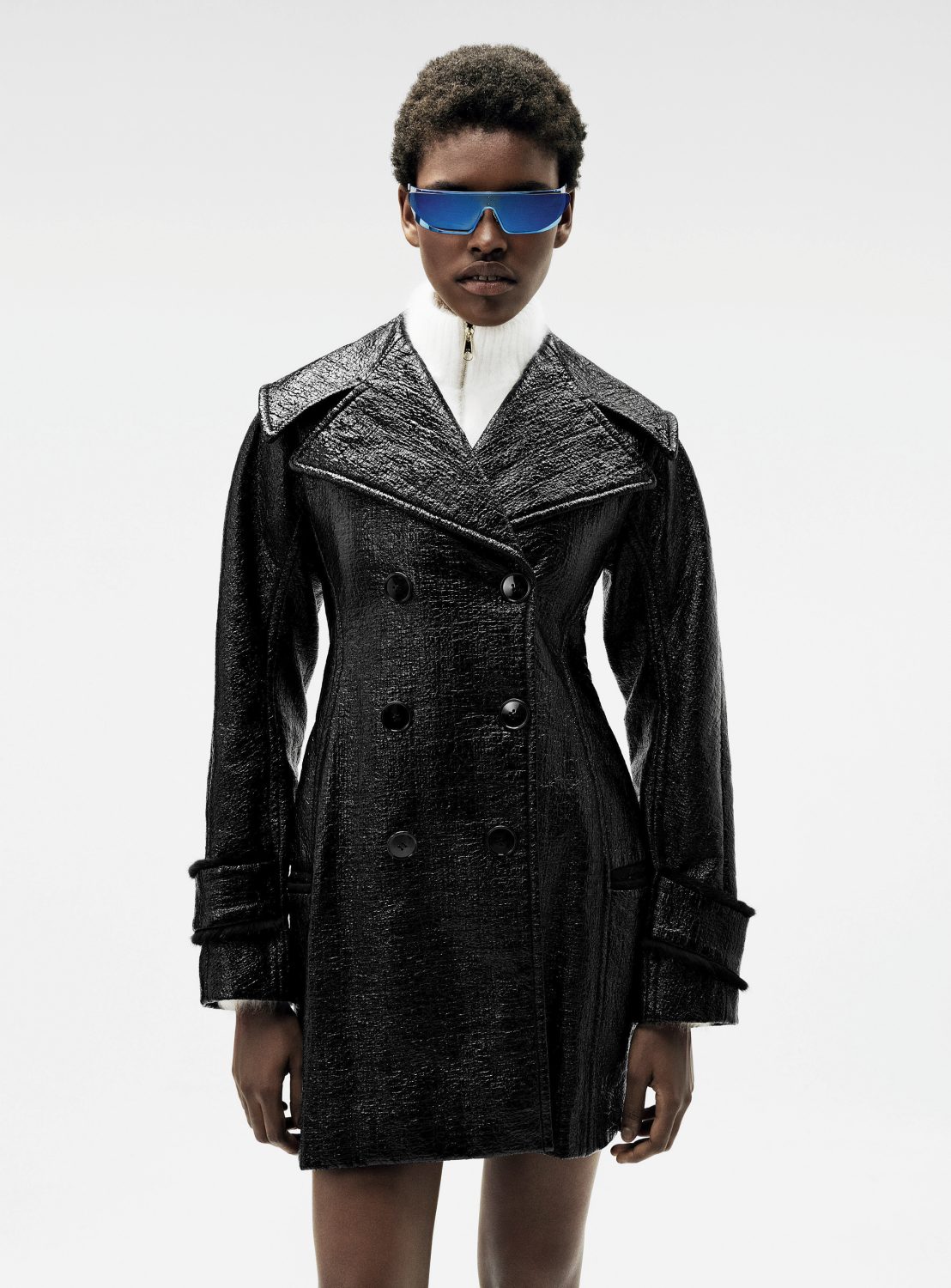 Woman in a shiny black trench coat and blue sunglasses in a studio. Fashion shoot with a modern, edgy style and clean white backdrop.