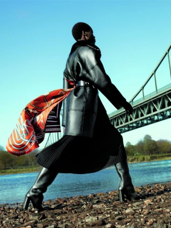 Model walking by a river under a steel bridge wearing a black leather coat and pleated skirt with an orange patterned scarf