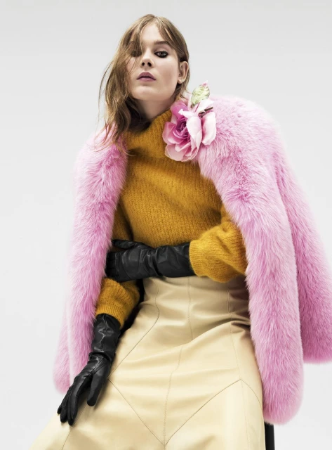 A stylish woman in a mustard sweater, tan skirt, and pink fur coat poses in a studio setting, exuding modern fashion and elegance.
