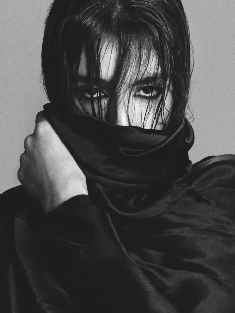 Monochrome portrait of a person with dark hair, styled forward, covering the lower face with a black silky fabric, intense gaze.