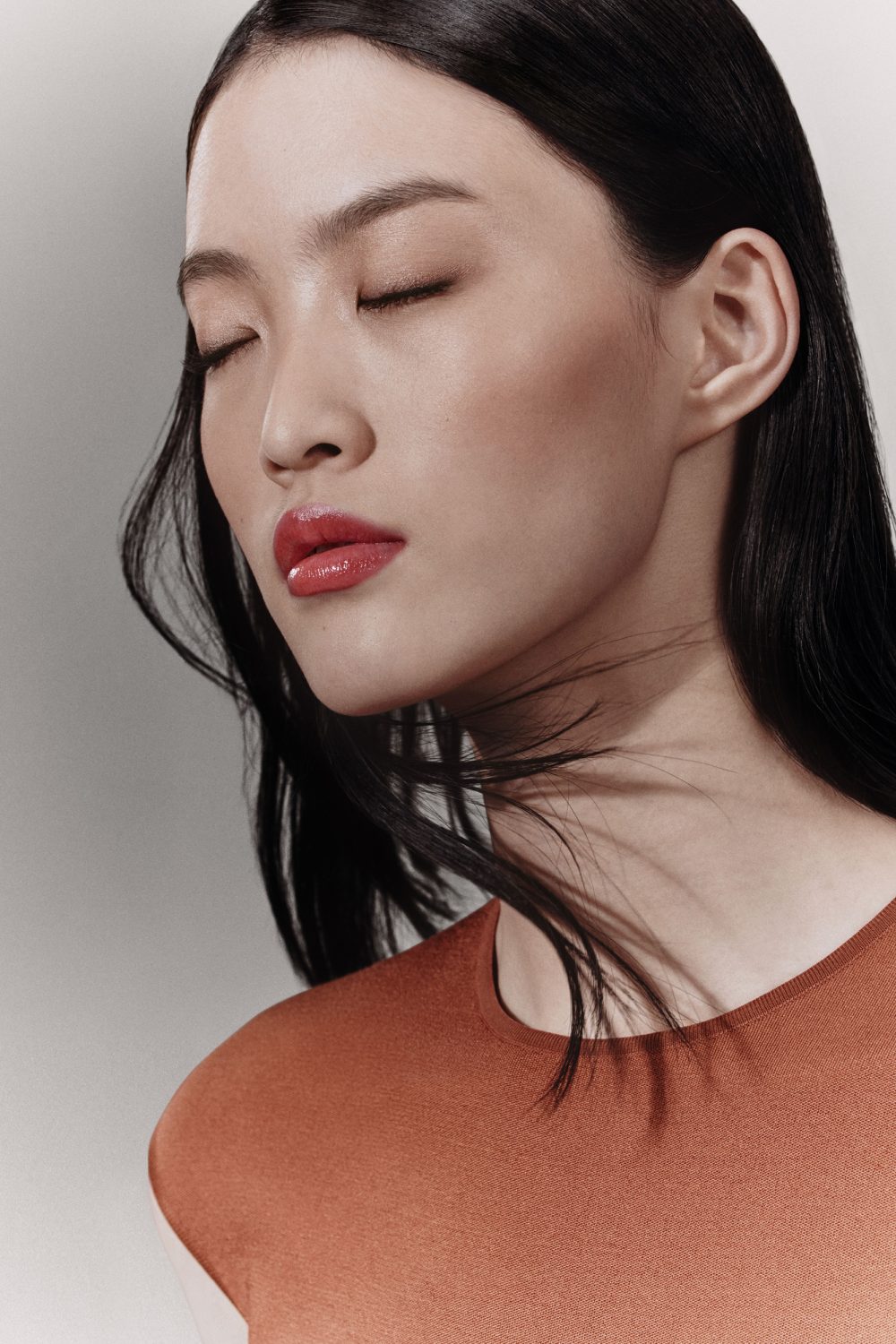 Close-up portrait of a woman with long dark hair and red lipstick wearing a burnt orange top against a neutral background.