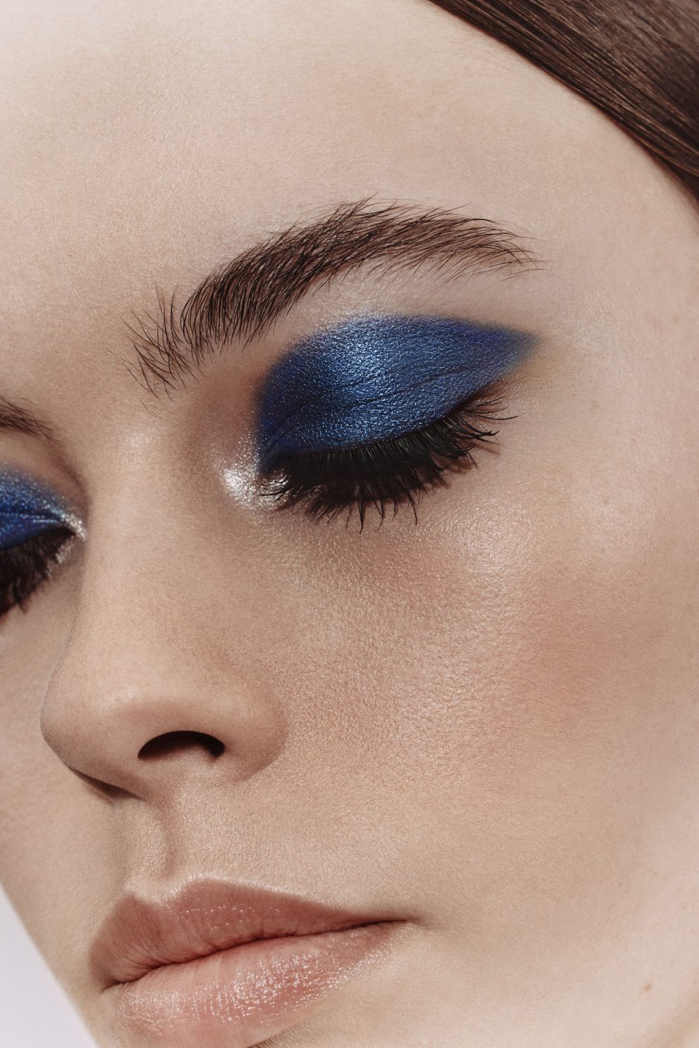 Close-up beauty portrait of a model with dramatic metallic blue eyeshadow and dark lashes against a neutral background.