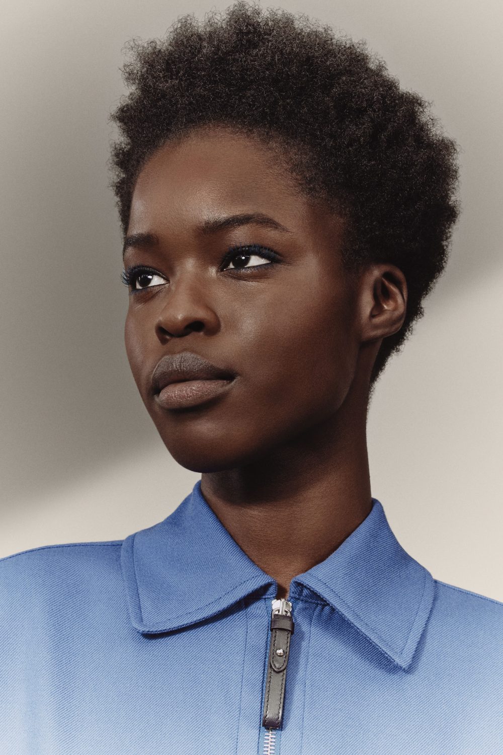 Close-up studio shot of an African American woman with short curly hair, wearing a blue collared shirt with a silver zipper.
