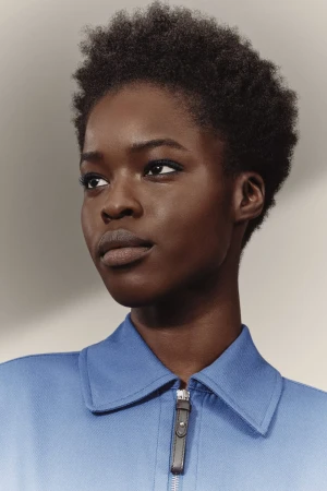 Close-up studio shot of an African American woman with short curly hair, wearing a blue collared shirt with a silver zipper.