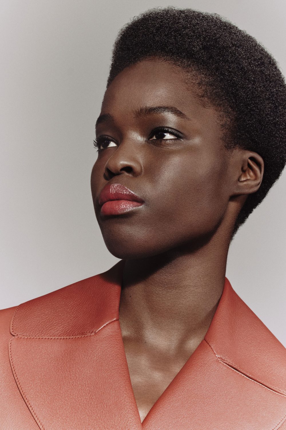 Close-up studio portrait of a young adult African woman with dark hair and an orange jacket, looking away from the camera.