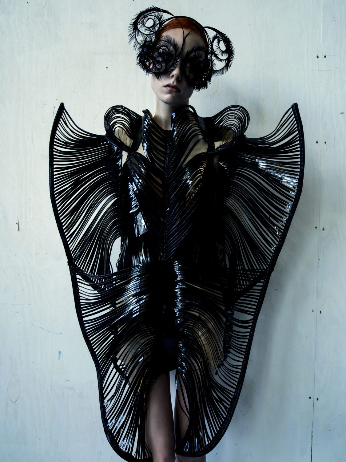 Fashion model wears a black, futuristic dress with linear accents and a feathered headpiece, posing against a light grey textured backdrop.