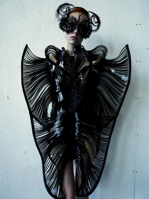 Fashion model wears a black, futuristic dress with linear accents and a feathered headpiece, posing against a light grey textured backdrop.