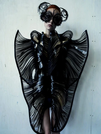 Fashion model wears a black, futuristic dress with linear accents and a feathered headpiece, posing against a light grey textured backdrop.