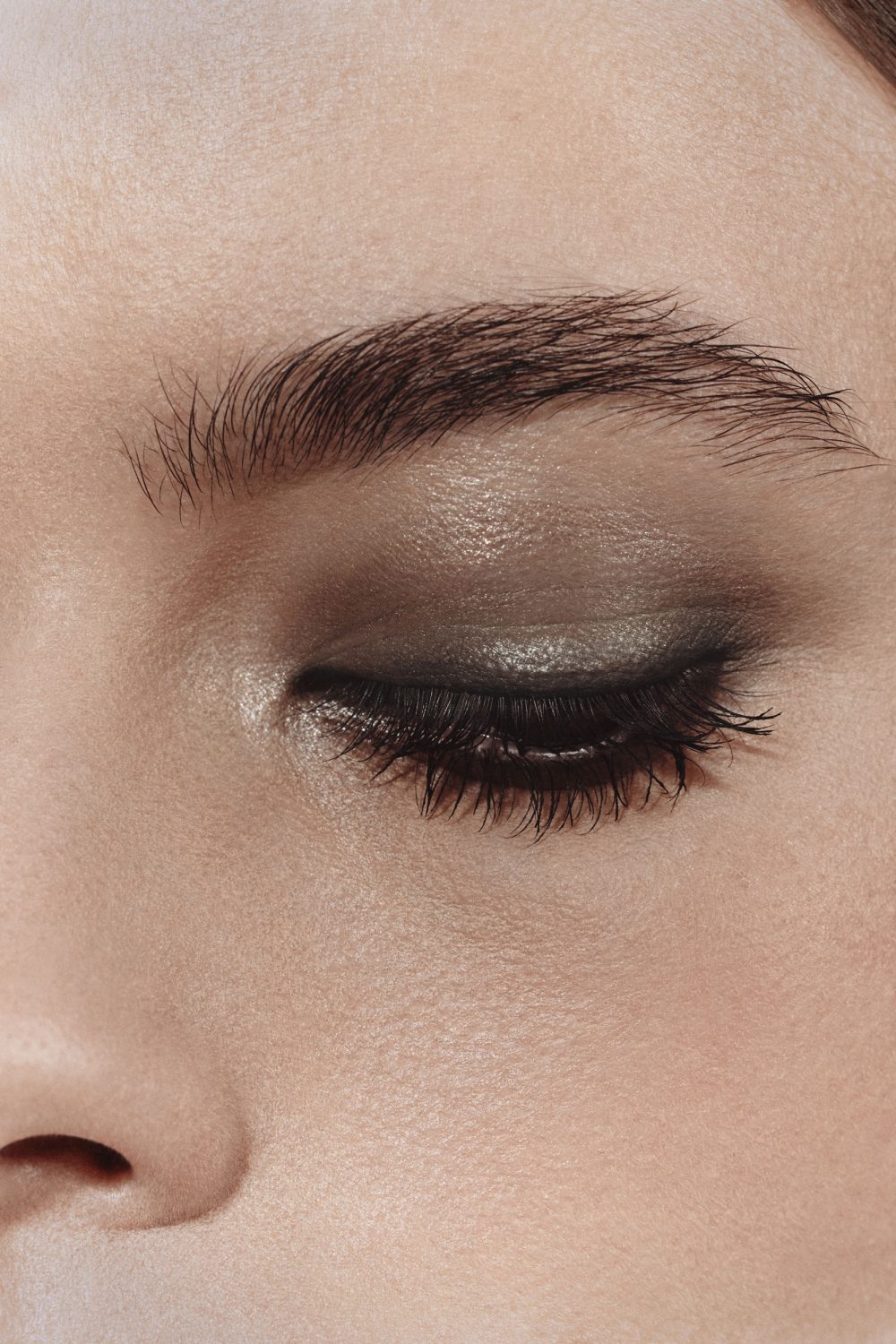 Close-up studio shot of a woman's closed eye featuring dark eye shadow, long eyelashes, and a neatly shaped eyebrow.