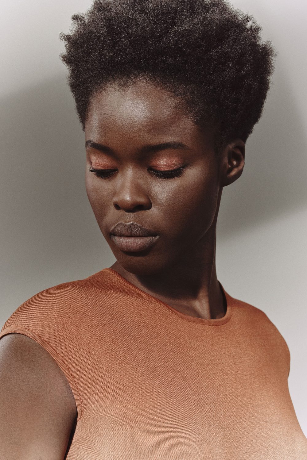 Portrait of an African American woman with copper eyeshadow and a copper top against a neutral background in a beauty editorial shot.