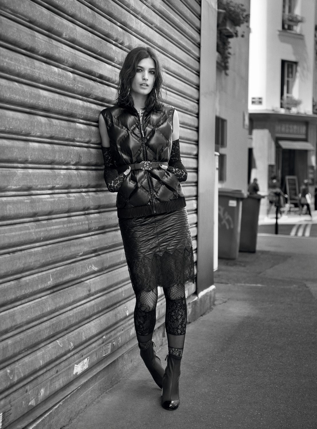 Monochrome fashion shot of a woman posing in an urban setting, wearing a puffer vest, skirt, lace leggings, and ankle boots, leaning on a wall.