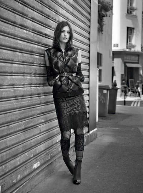 Monochrome fashion shot of a woman posing in an urban setting, wearing a puffer vest, skirt, lace leggings, and ankle boots, leaning on a wall.