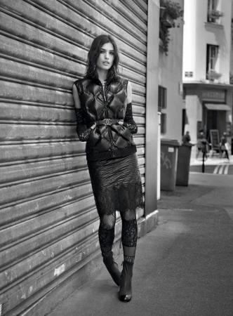 Monochrome fashion shot of a woman posing in an urban setting, wearing a puffer vest, skirt, lace leggings, and ankle boots, leaning on a wall.