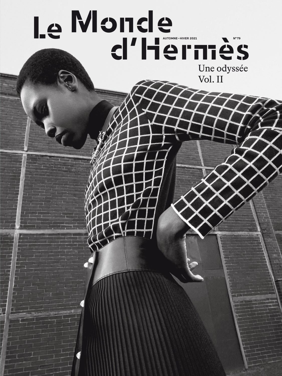 Fashion editorial shot of an African female model wearing a grid-patterned blouse and pleated skirt with a brick wall background.