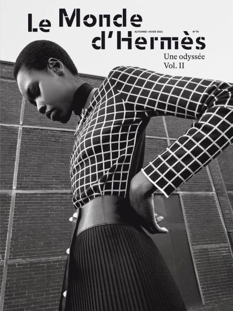 Fashion editorial shot of an African female model wearing a grid-patterned blouse and pleated skirt with a brick wall background.