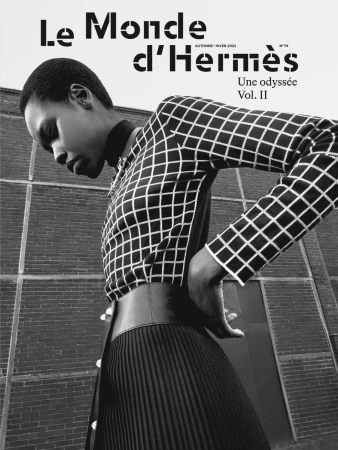 Fashion editorial shot of an African female model wearing a grid-patterned blouse and pleated skirt with a brick wall background.
