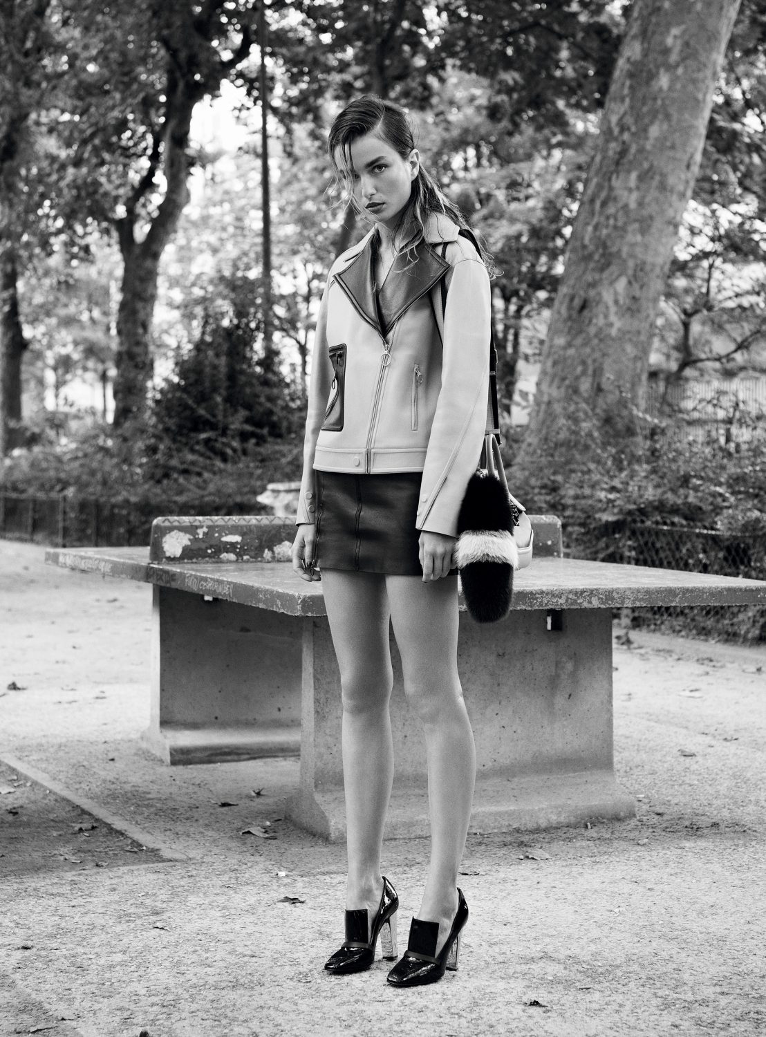 Black and white portrait of a model wearing a leather jacket and skirt standing outdoors near a park table.