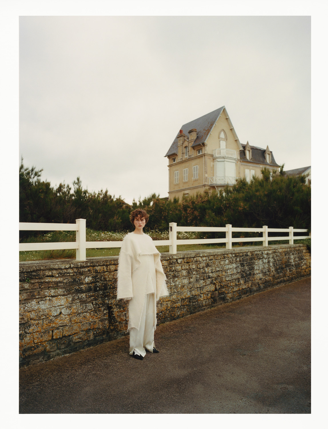 Woman in flowing white outfit stands on gray road in front of brick wall and large house under overcast sky. Fashion editorial shot.