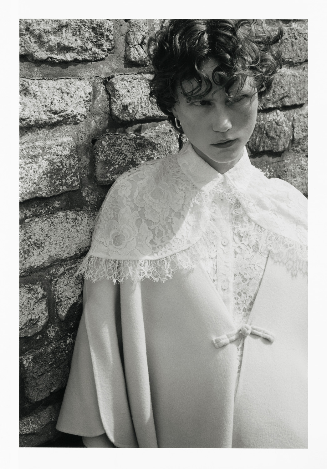 Black and white portrait of a model in a lace-trimmed cape in front of a stone wall, fashion photography, studio lighting