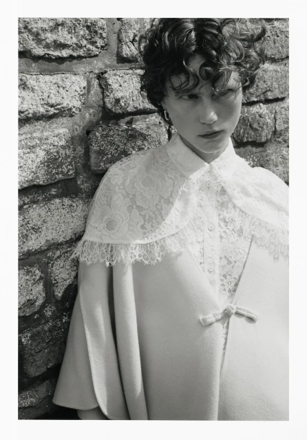Black and white portrait of a model in a lace-trimmed cape in front of a stone wall, fashion photography, studio lighting