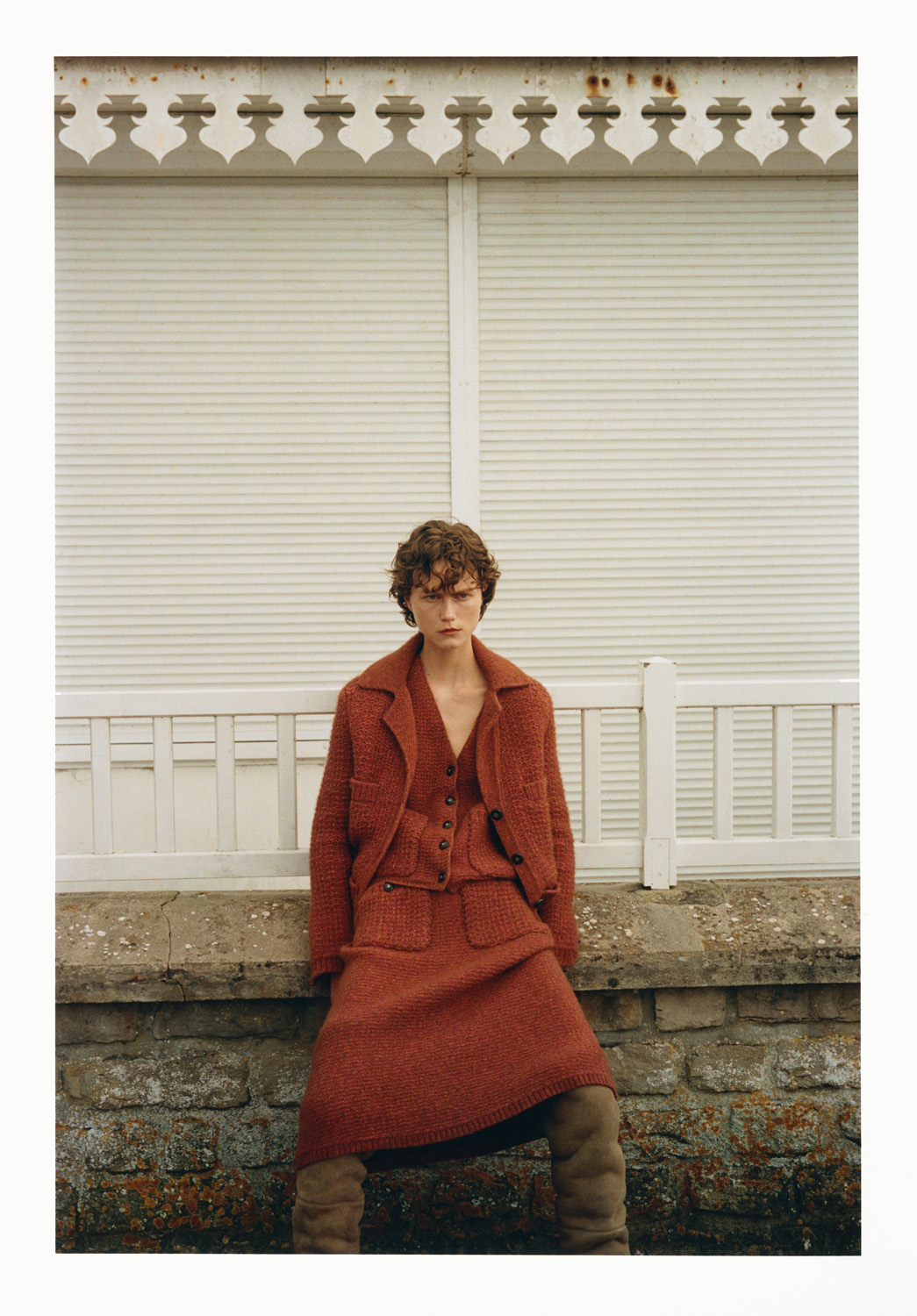 Fashion shot of a female model in a terracotta tweed jacket and skirt, posing against a weathered stone wall, with neutral-toned hosiery.