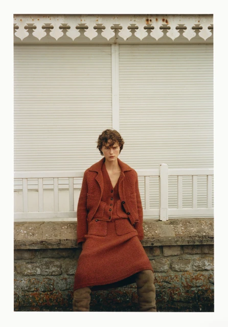 Fashion shot of a female model in a terracotta tweed jacket and skirt, posing against a weathered stone wall, with neutral-toned hosiery.
