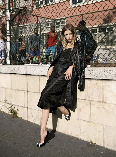 Stylish woman poses in a black dress and shiny coat by a park fence with basketball players in the background on a sunny day.
