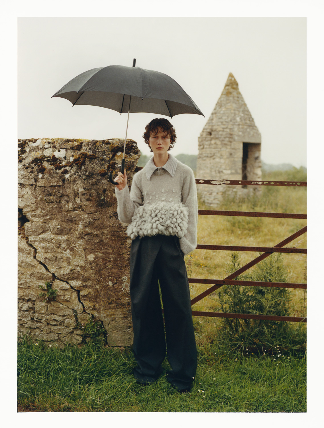 Editorial outdoor fashion shot of a young adult wearing a gray sweater and oversized trousers, holding an umbrella in a rural landscape.