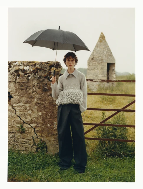 Editorial outdoor fashion shot of a young adult wearing a gray sweater and oversized trousers, holding an umbrella in a rural landscape.
