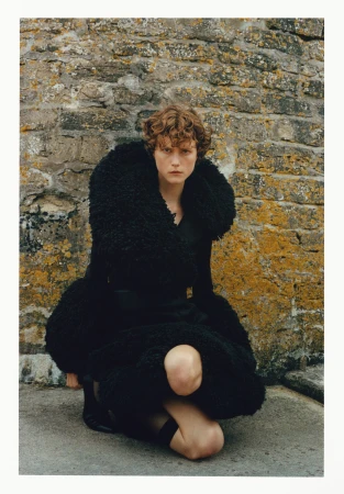 Model with curly hair crouching in a black textured coat against an old stone wall