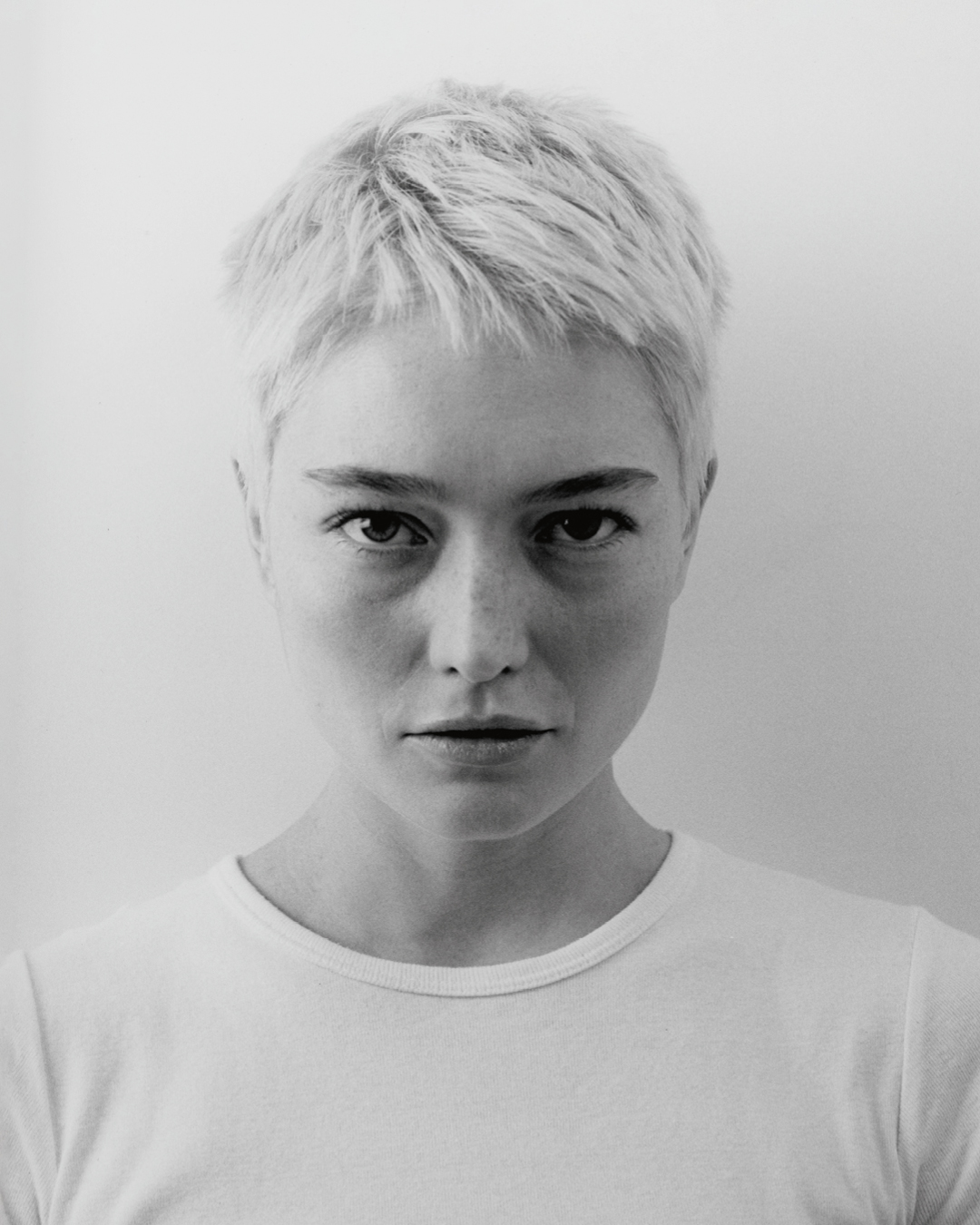 Close-up monochrome portrait of a woman with short blonde hair and a white t-shirt against a plain background
