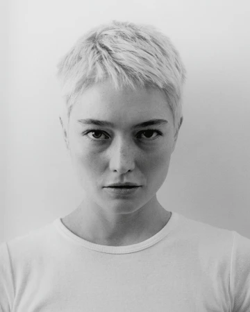 Close-up monochrome portrait of a woman with short blonde hair and a white t-shirt against a plain background