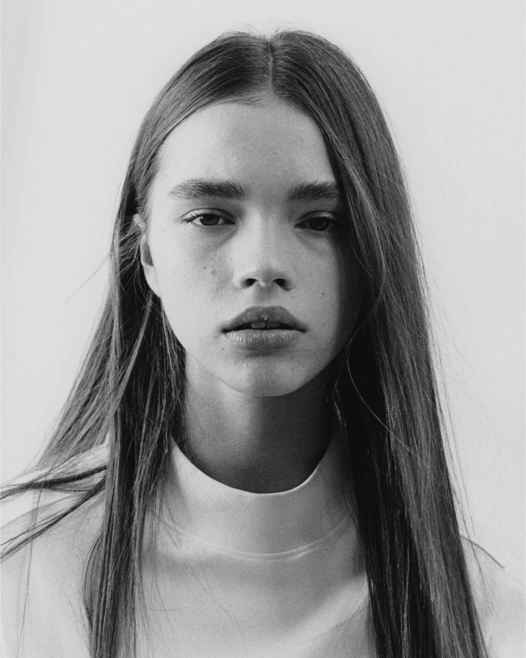 Monochrome portrait of a young woman with long straight hair, fair skin, and a neutral expression, wearing a white mock neck top, against a light background.